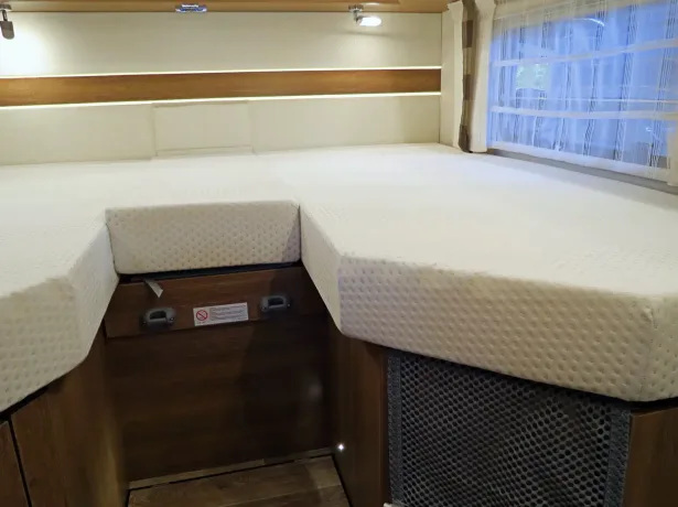 Heller Schlafbereich in einem Wohnmobil mit weißen Matratzen, Holzverkleidung, seitlichem Fenster mit Vorhängen und Leselampe.