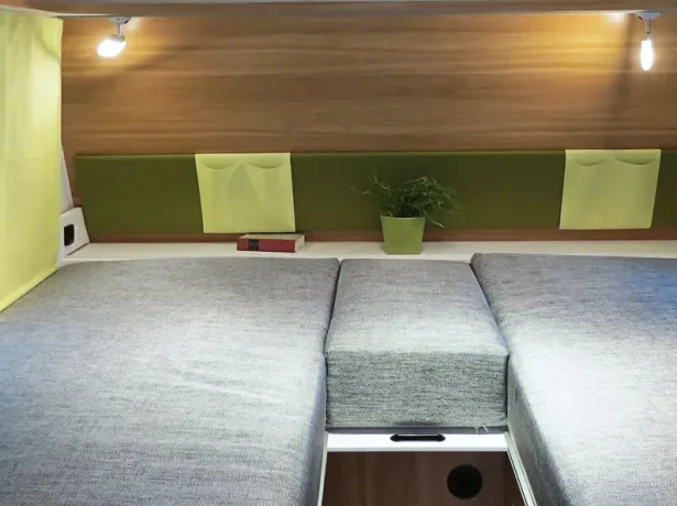 Modularer Schlafbereich mit grau-blauen Polstern, Holzrückwand, Leselampen, Aufbewahrungstaschen und einer Pflanze