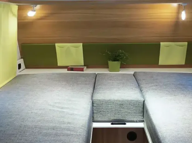 Modularer Schlafbereich mit grau-blauen Polstern, Holzrückwand, Leselampen, Aufbewahrungstaschen und einer Pflanze