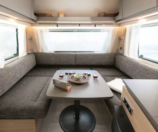 Wohnwagen-Innenraum mit U-förmiger braun- grauer Sitzgruppe, Klapptisch in der Mitte , großen Fenstern und hellen Oberschränken.