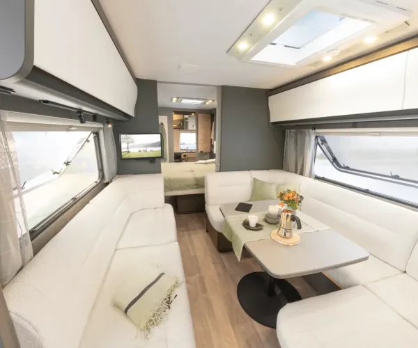 Modern eingerichteter Wohnwagen-Innenraum mit U-förmiger weißer Polstersitzgruppe, Klapptisch großen Fenstern und Dachluke.
