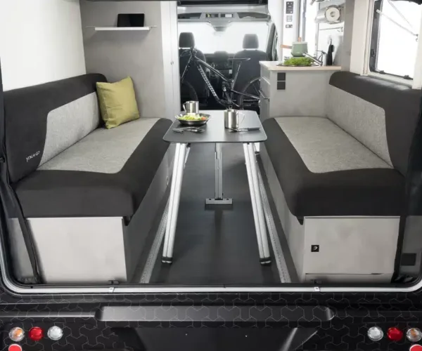 Blick in das Heck eines Wohnmobils mit zwei gegenüberliegenden Sitzbänken, klappbarem Tisch in der Mitte und Fahrrad im Hintergrund.