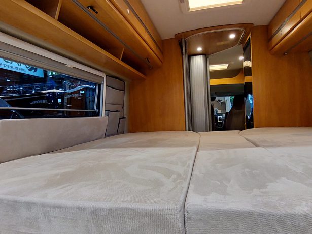 Schlafbereich in einem Camper Van mit beigen Matratzen, Holzverkleidung, seitlichen Fenstern und Hängeschränken.